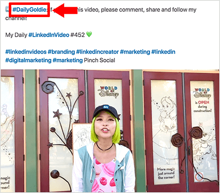 Ceci est une capture d'écran qui illustre comment Goldie Chan utilise des hashtags dans le texte de ses publications vidéo LinkedIn. Les légendes rouges pointent vers le hashtag #DailyGoldie dans le texte, qui est unique à ses publications vidéo et l'aide à suivre les partages. La publication comprend également d'autres hashtags pertinents qui aident les gens à trouver sa vidéo, y compris #LinkedInVideo. Dans l'image vidéo, Goldie se tient devant certaines portes lors d'un affichage de World of Disney. C’est une femme asiatique aux cheveux verts. Elle porte une casquette LinkedIn noire, un collier ras du cou noir, une chemise rose à imprimé macaron et une veste bleue et blanche.
