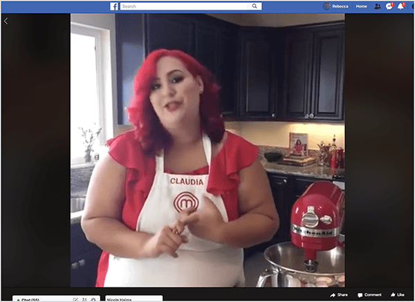 Voici une capture d'écran d'une vidéo Facebook Live mettant en vedette la chef Claudia Sandoval, qu'elle a animée en 2016 dans le cadre d'une promotion croisée avec l'application T-Mobile Tuesday. Dans la vidéo, Claudia se tient dans une cuisine avec des armoires noires et des comptoirs en granit. Une fenêtre au-dessus de l'évier laisse entrer la lumière naturelle dans la pièce. Claudia se tient à côté d'un mixeur Kitchen Aid rouge. C'est une femme latina qui a les cheveux rouge vif qui tombent juste en dessous de ses épaules. Elle porte du maquillage, un haut rouge vif et un tablier blanc avec Claudia et le logo MasterChef brodé en fil rouge. Pendant qu'elle parle, elle fait des gestes avec ses mains. En 2016, Stephanie Liu a collaboré avec Claudia pour produire cette vidéo en direct.