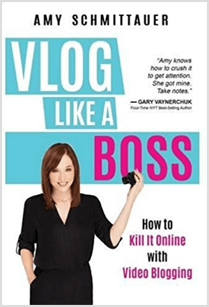 Amy Landino a écrit le livre Vlog Like a Boss sous le nom d'Amy Schmittauer. La couverture montre une photo d'Amy de la taille vers le haut tenant une caméra vidéo. Le titre apparaît sur un fond bleu clair avec des lettres blanches et fuchsia. Le slogan du livre est Comment le tuer en ligne avec les blogs vidéo.